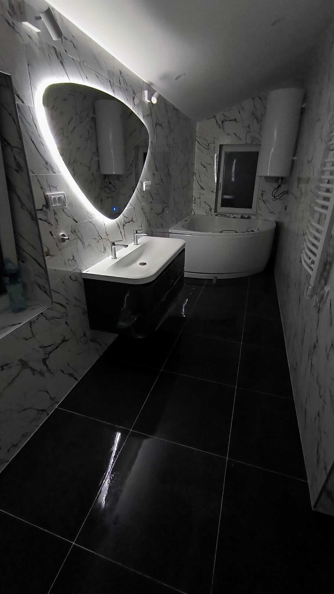 Schwarze Bodenfliesen, weiße Wandfliesen mit schwarzen Ziermustern, Eckbadewanne, längliches weißes Waschbecken mit zwei Wasserhähnen und schwarzem, schwebendem Unterschrank, großes Spiegel mit hintergrundbeleuchtetem weißen Licht und Touch-Schalter.