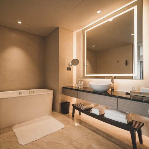 Decke mit kleinen runden Leuchten, moderne weiße Badewanne, grau-beige Wände, gleiche Fliesen auf dem Boden mit dunklen Ziermustern. Kleines rundes Waschbecken auf einer breiten schwarzen Marmorplatte mit weißen Ziermustern und grauen Schubladen, darüber ein großes quadratisches Spiegel mit Hintergrundbeleuchtung.